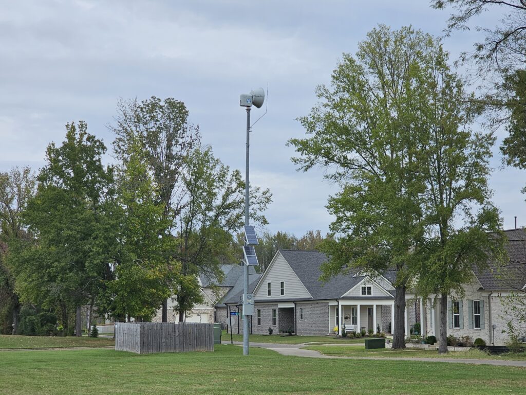 Solar-powered outdoor warning siren on a tall metal pole in a grassy Rossville neighborhood, with trees and homes under an overcast sky.
