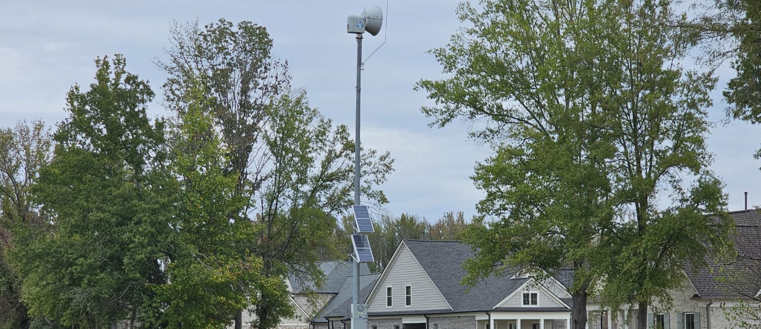 Solar-powered outdoor warning siren on a tall metal pole in a grassy Rossville neighborhood, with trees and homes under an overcast sky.