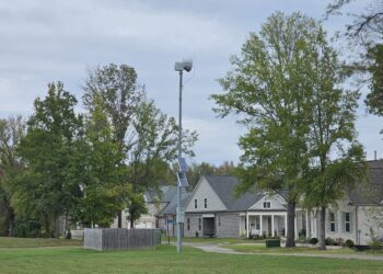 Solar-powered outdoor warning siren on a tall metal pole in a grassy Rossville neighborhood, with trees and homes under an overcast sky.